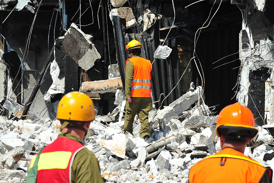 Emergency response personnel in protective gear assessing the scene of a collapsed building amid concrete debris and structural damage.