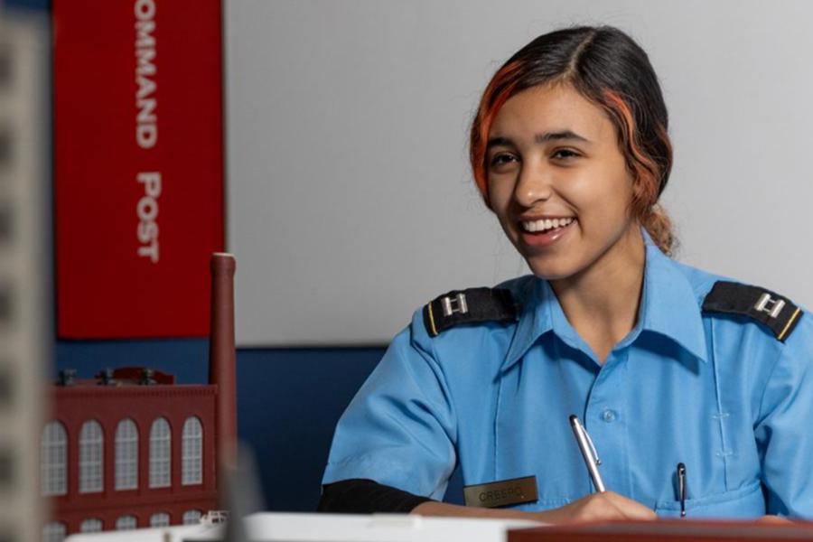 A smiling young woman in a light blue short-sleeed uniform shirt sits at a desk holding a pen. She has dark hair with a distinct orange streak framing her face, and her uniform features dark shoulder boards with captain's bars insignia.