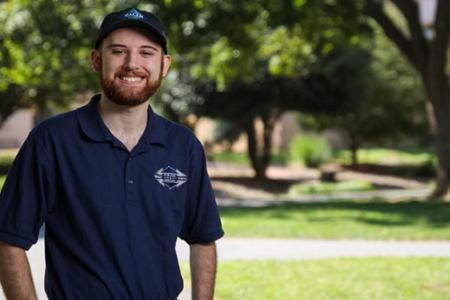 A smiling young man with a red beard standing outdoors wears a dark blue polo shirt and a matching baseball cap, both featuring a small logo. He stands on the left side of the frame against a blurred background of green grass and trees.