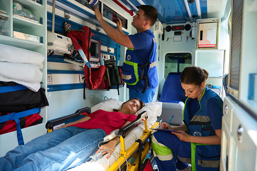 Paramedics providing care to a patient inside an ambulance, with one clinician preparing equipment and another documenting patient information.