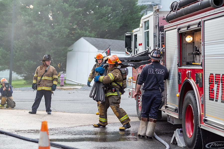 A group of firefighters in full turnout gear work near a red and white fire engine. One firefighter carries a bundled hose on their shoulder while another person wearing a dark shirt labeled 