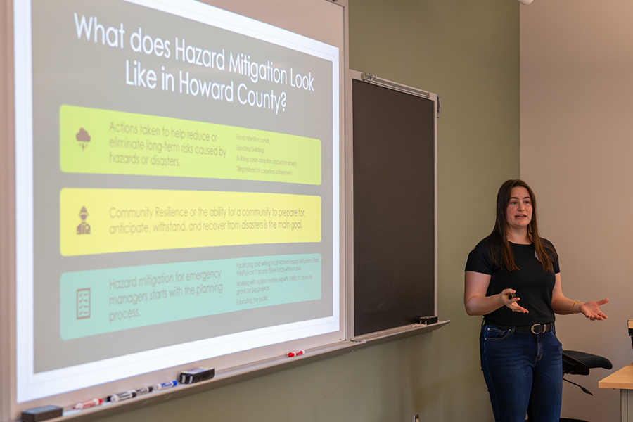 A woman stands in a classroom giving a presentation on Hazard Mitigation in Howard County. She gestures toward a projection screen displaying definitions of community resilience and emergency planning next to a large chalkboard.