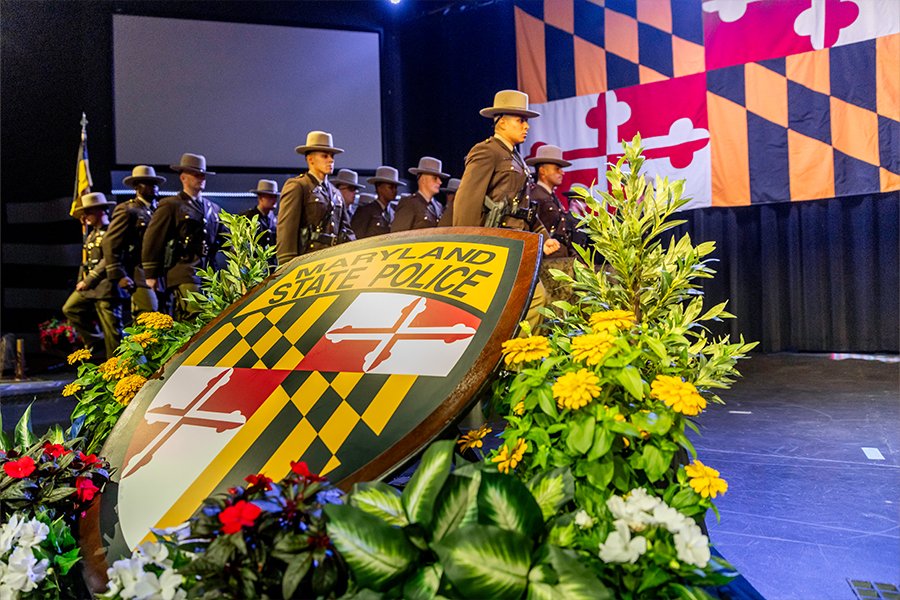 A large Maryland State Police shield emblem sits amid floral arrangements on a stage, with a line of state troopers marching in uniform and the Maryland state flag visible in the background.