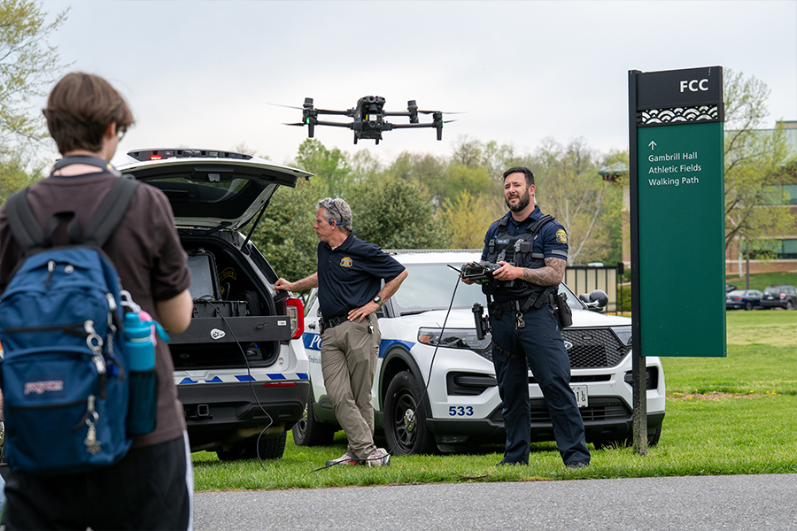 Law enforcement officers demonstrating drone technology on a college campus, with a drone in flight, police vehicles, and students observing nearby.