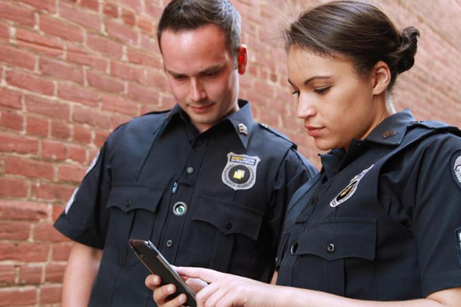 Two public safety officers in uniform reviewing information on a mobile device while standing outdoors near a brick wall.