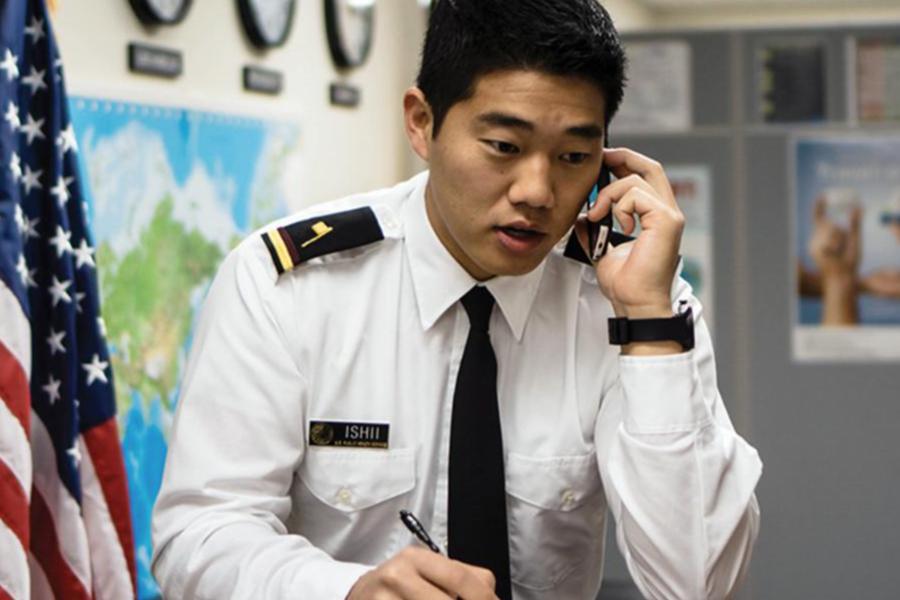 Emergency management professional speaking on a mobile phone while taking notes in an office, with maps and emergency planning resources visible in the background.