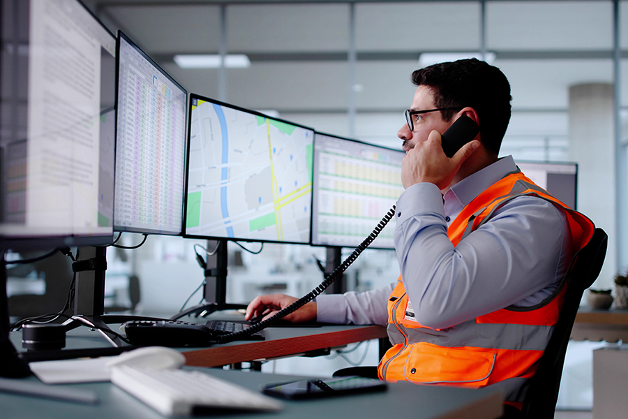 A man wearing glasses and a bright orange safety vest sits at a desk, speaking on a telephone receiver while looking at multiple large computer monitors displaying maps, spreadsheets, and data documents in an office or dispatch center environment.