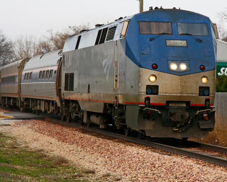 A passenger train travels along railroad tracks, led by a blue-and-silver locomotive with several passenger cars following behind.
