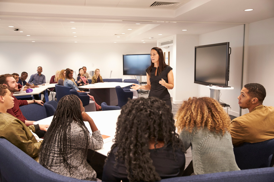 A woman stands at the center of a modern training room, speaking to a diverse group of participants seated around tables arranged for discussion and collaboration. Attendees face the presenter and appear engaged in the session. Large display screens and comfortable seating are visible throughout the room, suggesting a workshop, seminar, team meeting, or professional development event.