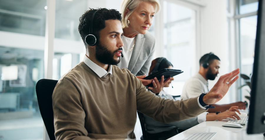 A customer service representative wearing a headset works at a computer while a supervisor stands nearby offering guidance. Other employees wearing headsets work at computer stations in the background.