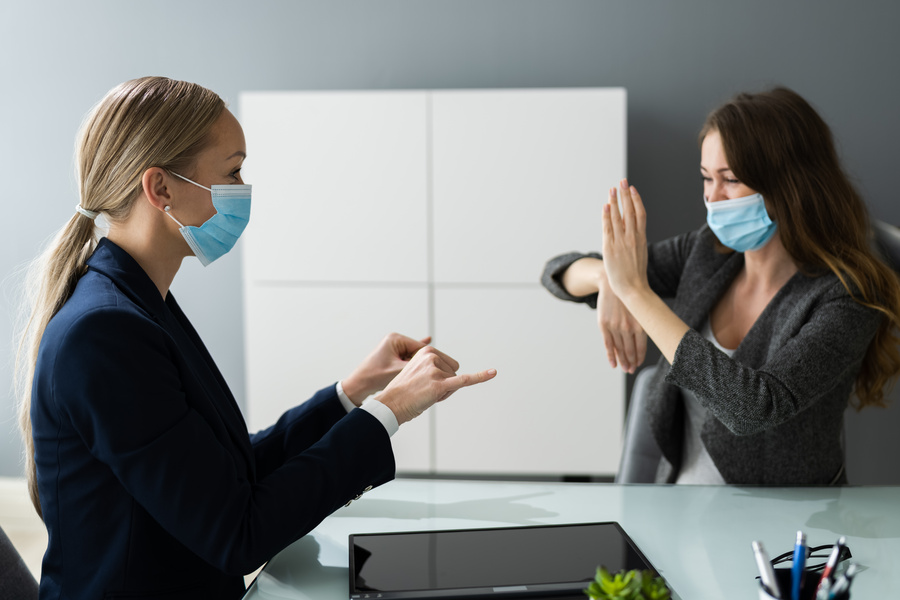 Two women wearing face masks sit across from each other at a desk in an office. One woman appears to be using hand gestures while communicating with the other, who is responding with her hands raised. A tablet and office supplies are visible on the desk, and a whiteboard stands in the background.