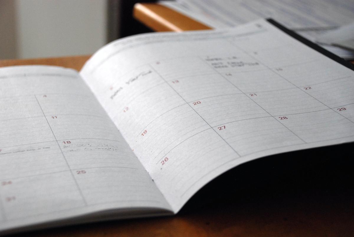 An open, black-backed monthly planner sits on a wooden desk, showing blank calendar grids with faint handwritten notes.