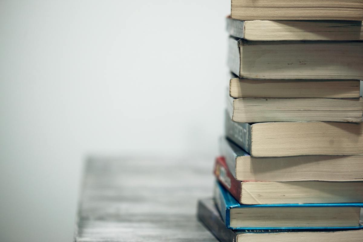 A neat stack of hardback and paperback books sits on the edge of a rustic wooden surface against a plain white background.