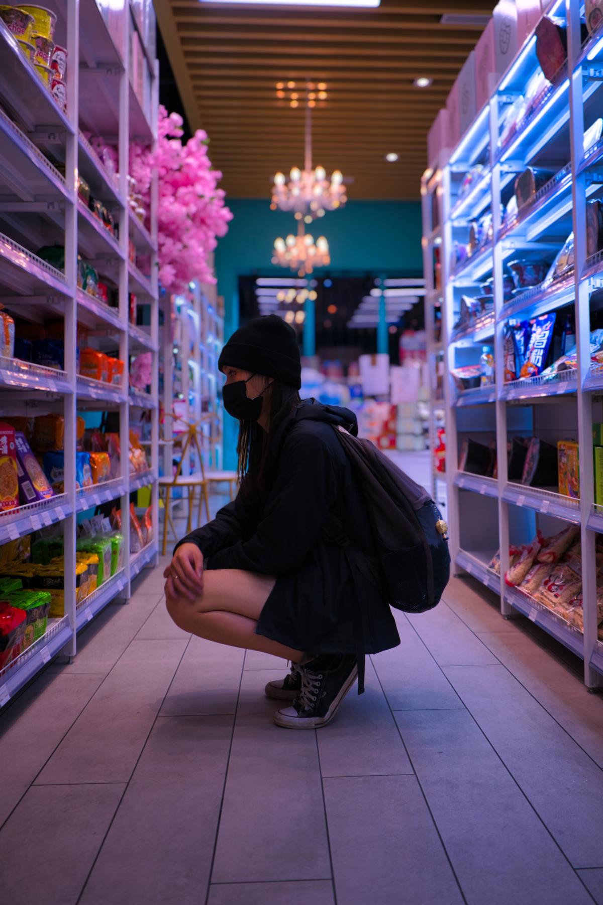 A person wearing a black beanie, face mask, and backpack squats down in a brightly lit store aisle filled with snacks, underneath a decorative chandelier and pink cherry blossom decorations.