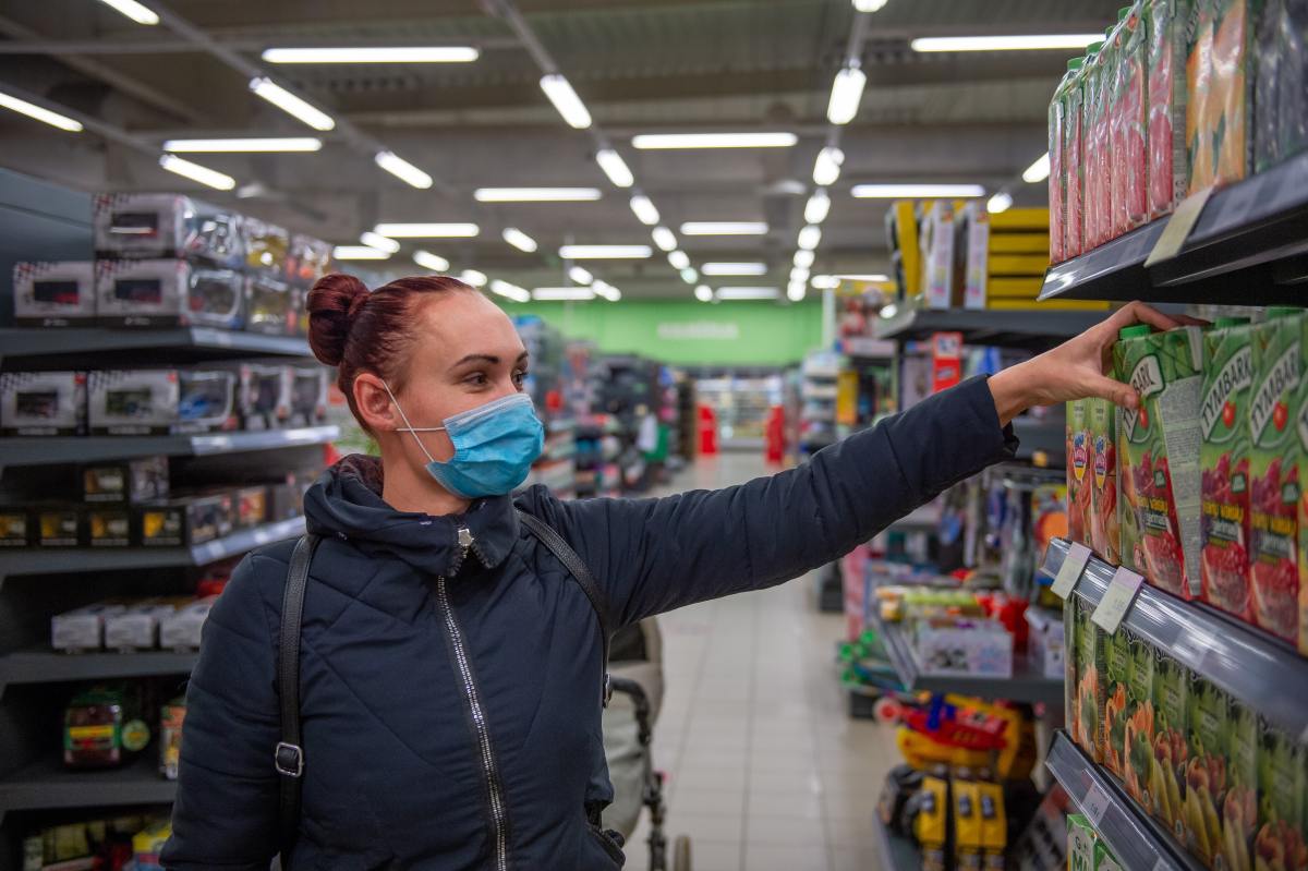 A woman wearing a blue face mask and a dark winter jacket reaches for a carton of juice on a grocery store shelf.