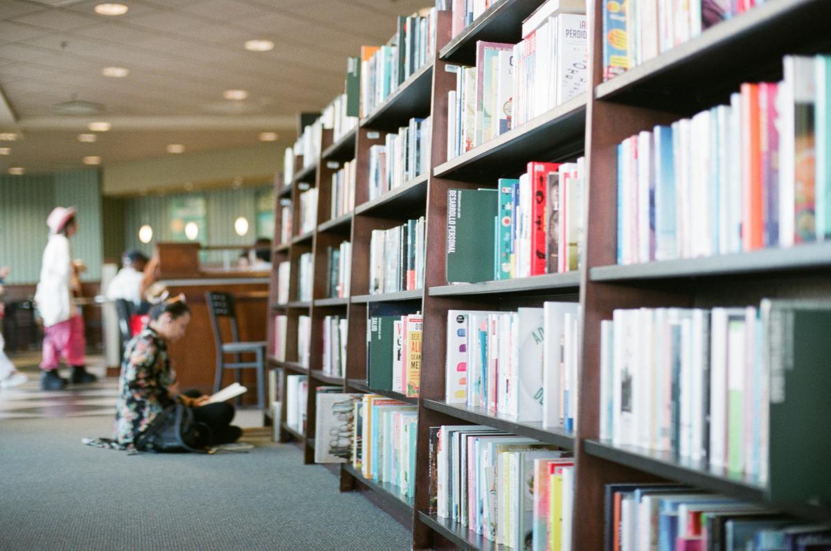 Tall, dark wooden bookshelves filled with colorful books line a brightly lit library aisle where a patron sits cross-legged on the floor reading a book in the blurred background.