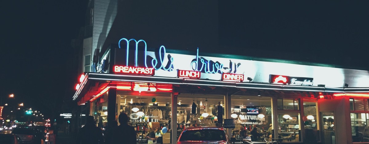 A classic retro diner exterior glows at night with blue and red neon signs reading