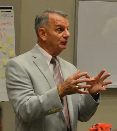 A man in a light grey suit jacket and a striped tie gestures with both hands while speaking in a classroom or office setting.